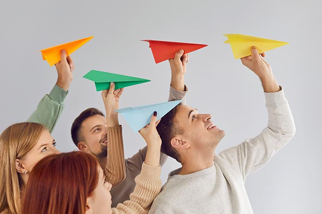 Joyful group of young people enthusiastically launching colorful paper airplanes into air.